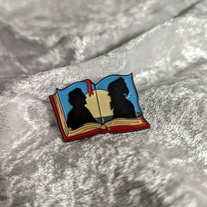 May include: A colorful enamel pin in the shape of an open book. The pin features black silhouettes of two people on the blue pages, with a yellow center and a red bookmark. The book has a red cover and a gold border.
