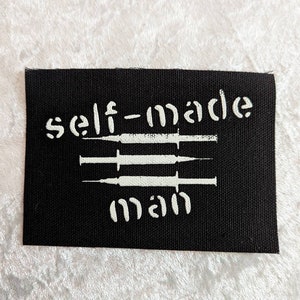 Puede incluir: Parche de tela negro con texto blanco que dice "self-made man" y tres jeringas blancas.