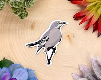 May include: A sticker featuring a gray and white bird with a black eye. The bird is perched on a branch with its wings spread out.
