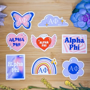 Könnte beinhalten: Ein Set aus neun Aufklebern mit verschiedenen Designs mit den Buchstaben "AΦ" und den Worten "Alpha Phi". Die Aufkleber befinden sich auf einer Holzoberfläche. Die Designs umfassen einen Schmetterling, eine Wolke, ein Herz mit Flügeln, einen Regenbogen, eine Blume und ein Wirbelmuster.
