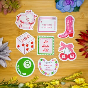 May include: A set of nine stickers featuring various designs related to the sorority Pi Beta Phi. The stickers are on a wooden surface and include a heart wreath, a computer screen with the text "Pi Beta Phi is so fetch", a dice with the letters "ПВФ", a cherry with the numbers 18 and 67, a green 8-ball, a heart with the text "Love Pi Beta Phi is my lucky charm", and a cowboy boot with the letters "ПВФ".