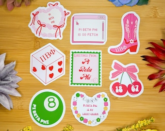 May include: A set of nine stickers featuring various designs related to the sorority Pi Beta Phi. The stickers are on a wooden surface and include a heart wreath, a computer screen with the text "Pi Beta Phi is so fetch", a dice with the letters "ПВФ", a cherry with the numbers 18 and 67, a green 8-ball, a heart with the text "Love Pi Beta Phi is my lucky charm", and a cowboy boot with the letters "ПВФ".