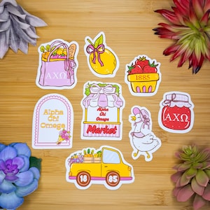 May include: A collection of colorful stickers featuring various designs. Included are a pink tote bag with the letters AXΩ, a yellow lemon, a basket of strawberries, a market stand, a duck, a jar of jam, and a yellow truck.