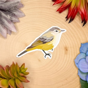 May include: A sticker featuring a yellow and brown bird with a white belly. The bird is perched on a light-colored wooden surface, surrounded by colorful artificial flowers. The bird sticker is a nature-themed item.