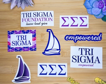 May include: A collection of stickers featuring the Tri Sigma sorority logo and the phrase "empowered". The stickers are in various colours and designs, including a sailboat, a geometric pattern, and a simple text design. The stickers are on a wooden surface.