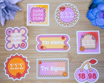 May include: Eight colourful stickers with various designs featuring the Greek letters Sigma Sigma Sigma (ΣΣΣ) and the words "Tri Sigma". The stickers are on a wooden surface.