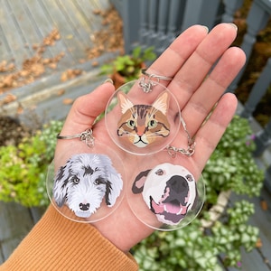 May include: Three round clear acrylic keychains with different animal faces. The keychains feature a tabby cat, a white dog, and a gray and white dog. The keychains are held in a person's hand.