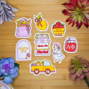 Könnte beinhalten: Eine Sammlung farbenfroher Aufkleber mit verschiedenen Designs. Enthält eine rosa Tragetasche mit den Buchstaben "AΦ", eine gelbe Zitrone mit rosa Schleife, einen Korb mit Erdbeeren und einen Marktstand mit dem Text "Alpha Phi Market".