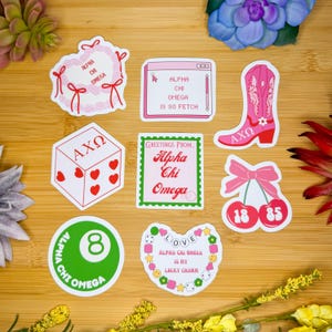 Può includere: Un set di nove adesivi con una varietà di design, tra cui una ghirlanda di cuori, uno schermo del computer, uno stivale da cowboy, un dado, una ciliegia, una palla da biliardo e un ciondolo a forma di cuore. Gli adesivi sono tutti rosa e verdi e presentano il testo "Alpha Chi Omega".