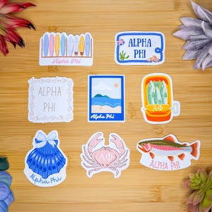 Könnte beinhalten: Satz von neun Aufklebern mit verschiedenen Strandmotiven mit dem Text "Alpha Phi". Die Aufkleber beinhalten einen Surfboardständer, eine Muschel, eine Krabbe, einen Fisch, eine Strandszene, einen Fischmarkt und ein Schild mit einem Seilrand.