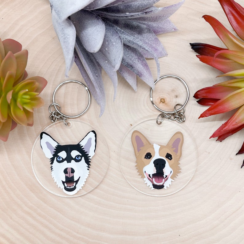 May include: Two round keychains with dog faces on them. The keychain on the left has a black and white husky face. The keychain on the right has a brown and white corgi face.