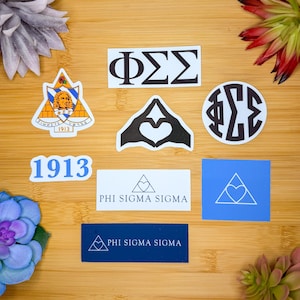 May include: A collection of stickers and decals featuring Greek letters, hearts, and the text "PHI SIGMA SIGMA" and "1913". The stickers are in various shapes and colors, including white, blue, and navy, and are arranged on a wooden surface.