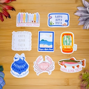 May include: A collection of Kappa Alpha Theta stickers featuring various designs. Includes stickers with surfboards, a nautical theme, a seashell, a crab, a fish, and a can of fish. The stickers are in various colors, including blue, pink, and yellow.