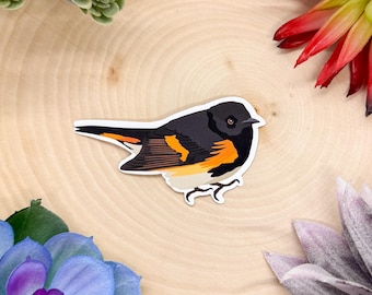 May include: A black and orange bird sticker with a white background. The bird has a black head, a white belly, and orange and black wings.