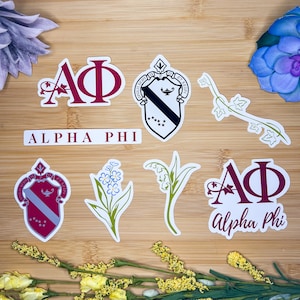 Könnte beinhalten: Ein Set mit sieben Aufklebern der Alpha Phi Sorority. Die Aufkleber zeigen das Wappen der Sorority, die Buchstaben "APhi" und florale Designs. Die Aufkleber sind auf weißem Vinyl gedruckt und haben eine glänzende Oberfläche.