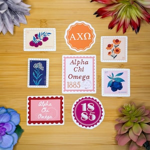 May include: A collection of decorative stickers with various designs, including floral illustrations, the Greek letters AXΩ, and the words "Alpha Chi Omega 1885". The stickers are arranged on a wooden surface.