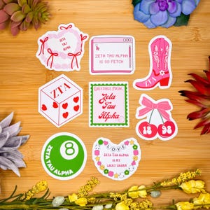 May include: Nine colorful stickers featuring the Greek letters Zeta Tau Alpha. The stickers include a heart with a ribbon, a computer screen, a dice, a cherry, a heart with a charm, and a circle with the number 8. The stickers are on a wooden surface.