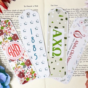 May include: Four colorful bookmarks with different designs. One bookmark features a floral pattern with a red monogram, another has a white background with blue stars and numbers, a third has a green background with white polka dots and the letters AXQ, and the last bookmark has a floral pattern with a red and white harp and the text "Alpha Chi Omega, Real Strong Women".
