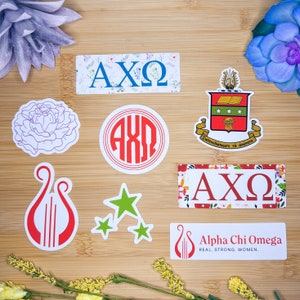 May include: A set of eight stickers featuring the Greek letters AXΩ, a red and white lyre, a green star, a red circle with the letters AXΩ, a floral design with the letters AXΩ, a crest with the letters AXΩ, and a floral design with the text "Alpha Chi Omega REAL. STRONG. WOMEN."