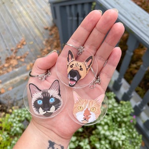 May include: Three clear round keychains with images of a cat, a dog, and a cat. The keychains are held in a hand.