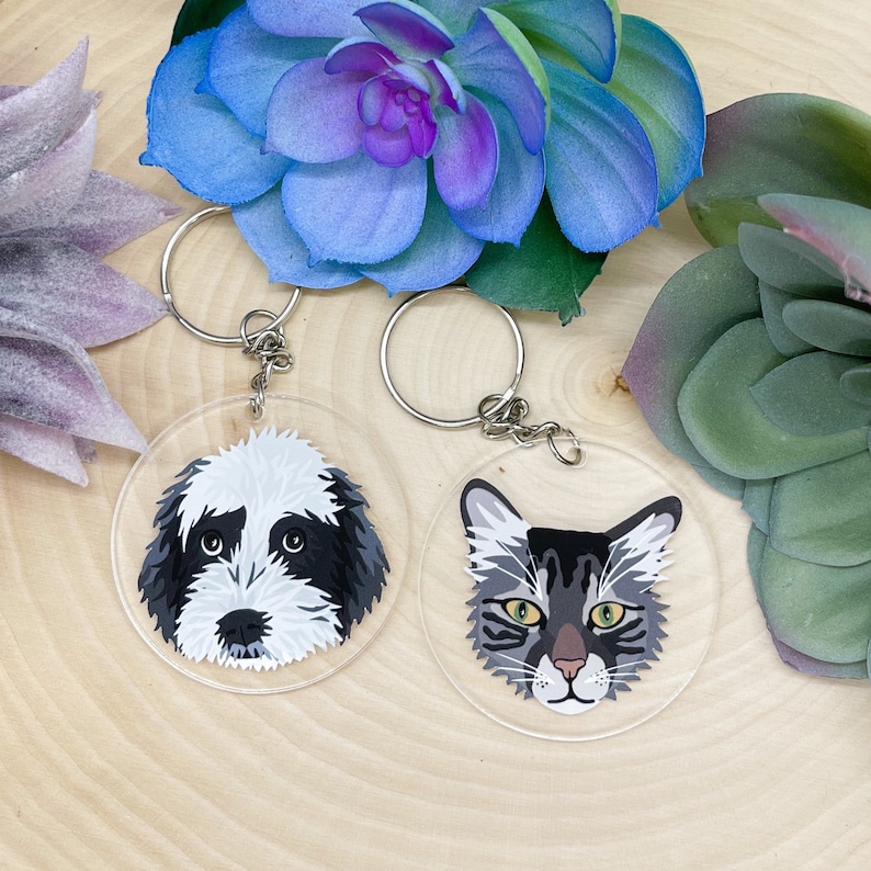 May include: Two round keychains with clear acrylic backgrounds. One keychain features a black and white illustration of a dog's face. The other keychain features a black and white illustration of a cat's face.
