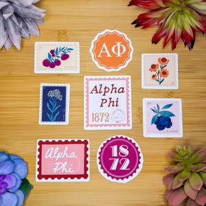 Könnte beinhalten: Eine Sammlung dekorativer Aufkleber mit verschiedenen Designs. Die Aufkleber enthalten Blumen- und Fruchtdarstellungen sowie den Text "Alpha Phi" und das Jahr "1872". Die Aufkleber sind in verschiedenen Formen und Farben gehalten.
