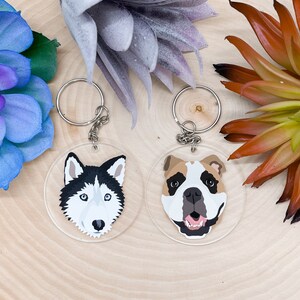 May include: Two round keychains with dog faces. The keychain on the left features a black and white husky with blue eyes. The keychain on the right features a brown and white bulldog with a pink tongue.