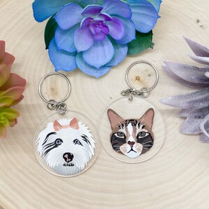 May include: Two round clear acrylic keychains with a white dog and a brown tabby cat printed on them. The keychains are hanging from silver key rings.