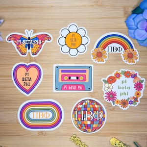 May include: A set of nine colorful stickers featuring a variety of designs, including a butterfly, a flower, a rainbow, a heart, a cassette tape, a wreath, a circle, and a disco ball. All stickers feature the text "Pi Beta Phi" in various styles and colors.