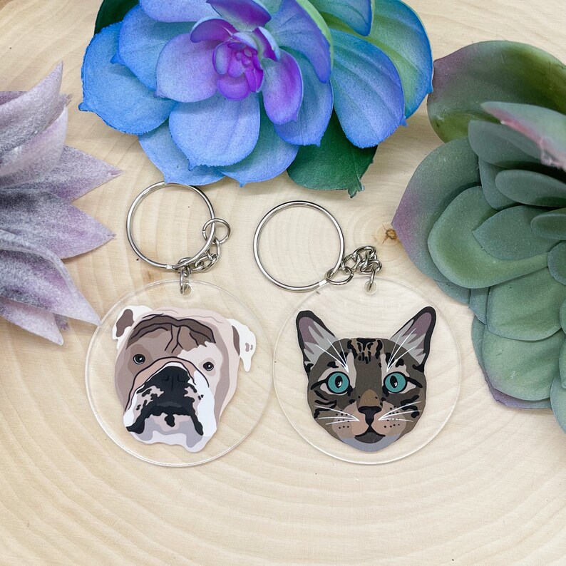 May include: Two clear acrylic keychains with a bulldog and a tabby cat printed on them. The keychains are on a wooden surface with two succulents.