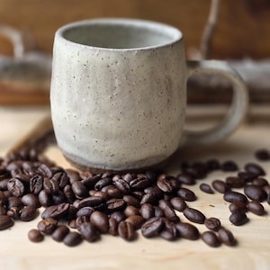 Puede incluir: Una taza de cerámica blanca con un esmalte moteado se encuentra sobre una superficie de madera rodeada de granos de café.