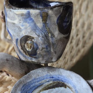 May include: A small, hand-painted ceramic cup and saucer set. The cup is blue, white and brown with abstract designs. The saucer is grey with a blue spiral design.