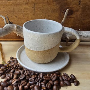 Op de afbeelding: Een witte en beige gespikkelde keramische mok met een handvat staat op een bijpassende schotel. Op de voorgrond ligt een stapel koffiebonen.
