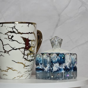 May include: A white ceramic mug with gold accents and a gold handle, featuring a black and gold marble design. Beside it is a decorative blue and white resin jar with a crystal-like lid.