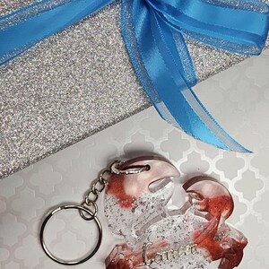 May include: A red and white resin keychain shaped like a crab with glitter embedded in the resin. The keychain has a silver metal ring and chain. The word "Cancer" is written in silver glitter on the crab's body.