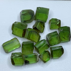 May include: A collection of small, loose, faceted green gemstones with a translucent appearance. The stones exhibit varying shades of vibrant olive and forest green, featuring sharp edges and polished surfaces that reflect light across their geometric shapes.
