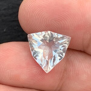 Può includere: Una gemma trasparente e sfaccettata a forma triangolare. La gemma presenta molteplici sfaccettature che riflettono la luce, creando un effetto scintillante. La pietra è tenuta tra le dita, mostrando la sua chiarezza e taglio.
