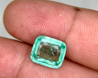 Puede incluir: Una gema verde esmeralda, tallada en forma cuadrada, con un aspecto ligeramente translúcido. La gema tiene forma rectangular con esquinas redondeadas, mostrando un color natural vibrante. La piedra se sostiene entre los dedos, resaltando su tamaño y claridad.
