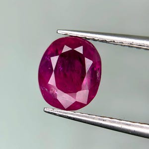 May include: A faceted, oval-shaped ruby gemstone held by tweezers. The ruby is a deep, vibrant red color with internal facets that reflect light. The tweezers are silver-colored and hold the gemstone against a light gray background.