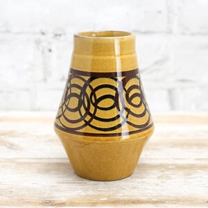 May include: A yellow ceramic vase with a brown geometric pattern. The vase has a wide base and a narrow neck.