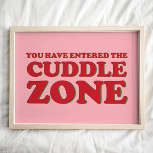 Op de afbeelding: Een ingelijste print met de tekst "YOU HAVE ENTERED THE CUDDLE ZONE" in rode letters met groene contouren op een roze achtergrond. De lijst is licht houtkleurig.