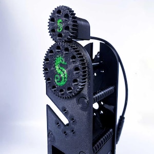 May include: Black mechanical device with two interlocking gears, one featuring a green seahorse design and the other a green number 5. The device has a complex structure with a screw and a black cable.