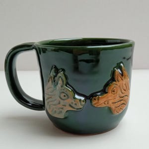 Puede incluir: Una taza de cerámica verde oscuro con un asa curvada. La taza presenta dos diseños de cabeza de lobo en relieve en tonos marrones y beige. La taza tiene un acabado brillante.