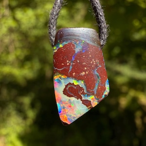 Peut inclure: Pendentif en opale aux couleurs vives, avec des nuances de rouge, de bleu et d'iridescent. Il est suspendu à un cordon gris foncé. La surface de la pierre précieuse présente un motif unique et une texture intéressante.