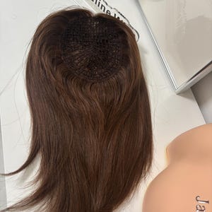 Pode incluir: Um topper de cabelo castanho com uma base de malha, projetado para adicionar volume e cobertura. A peça capilar é de cor castanho quente e parece ser feita de cabelo humano. O produto é exibido em uma superfície branca com o nome "Jacqueline" visível.