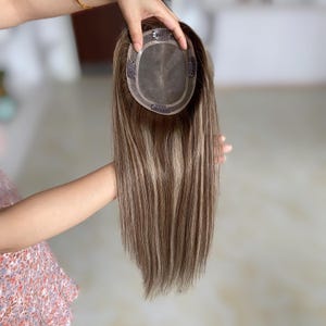 May include: A brown and blonde hair topper with a lace base and clips. The hair is long and straight, with highlights throughout. The topper is held up by a person.