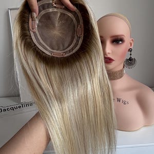 May include: A blonde hair topper with a brown base and clips. The hairpiece is held up, showcasing the cap and the long, straight blonde hair. A mannequin head is in the background.