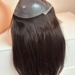 Op de afbeelding: Een donkerbruin haarstuk met een transparante basis wordt door een hand omhoog gehouden. Het haar is lang en steil, met een natuurlijke uitstraling. De basis lijkt gemaakt van een dun, flexibel materiaal.