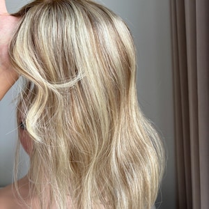 Puede incluir: Una peluca de cabello humano rubio con reflejos marrones claros. La peluca está diseñada con un estilo en capas y ondulado.