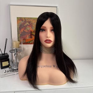 Op de afbeelding: Een mannequinhoofd met lang, steil zwart haar. De mannequin draagt een gouden choker en heeft een lichte huidskleur met rode lippenstift. De tekst "Jacqueline Wig" is zichtbaar op de borst van de mannequin.